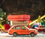 An orange toy car is going along a snow covered road against the background of festive lights and pines and carrying boxes with gifts. Concept of Christmas mood and preparation for the celebration