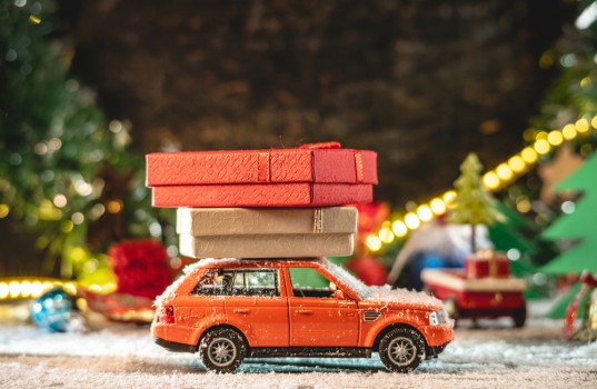 An orange toy car is going along a snow covered road against the background of festive lights and pines and carrying boxes with gifts. Concept of Christmas mood and preparation for the celebration