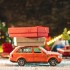 An orange toy car is going along a snow covered road against the background of festive lights and pines and carrying boxes with gifts. Concept of Christmas mood and preparation for the celebration